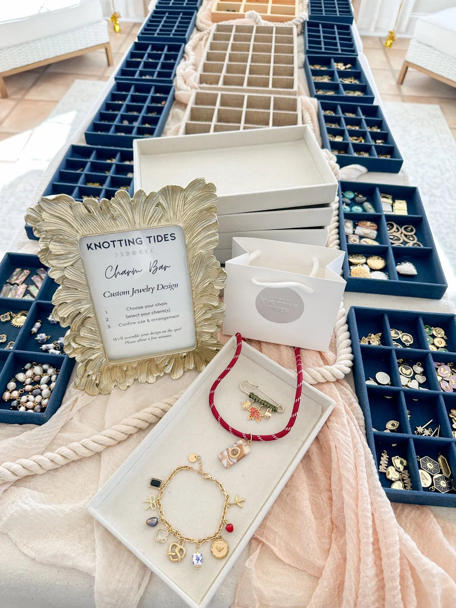 Knotting Tides Charm Bar table with navy charm trays, a gold framed sign and a finished charm bracelet