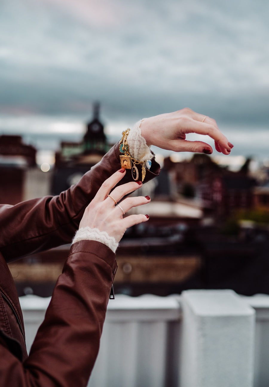 Hands showing a gold charm bracelet with a book locket against a city skyline